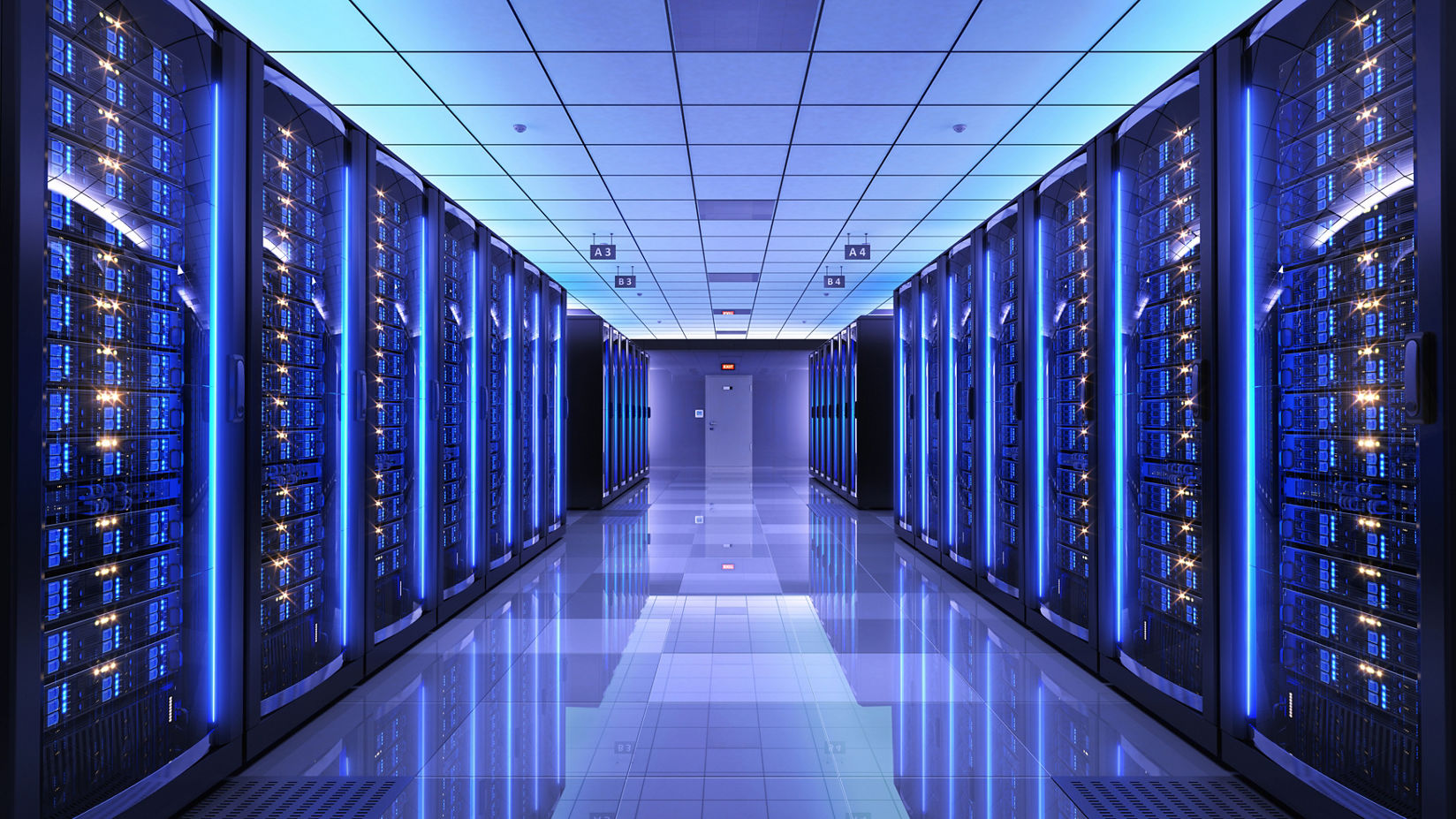 The view down a wide hallway in a server room. Server racks illuminated in blue light are shown on both sides of the walkway