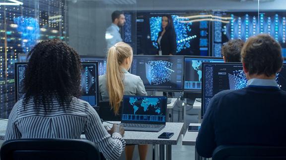 In a computer lab environment, two people stand at the front of the room having a conversation. Four other employees sit at PC workstations reviewing data models