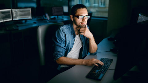 薄暗い作業エリアのデスクに座って、キーボードで入力しながらコンピューター画面の情報を確認する人