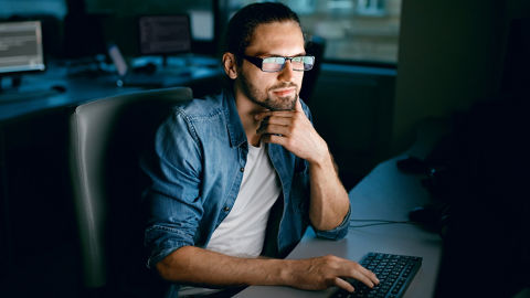 Person, die an einem Schreibtisch in einem schwach beleuchteten Arbeitsbereich sitzt und sich Informationen auf einem Computerbildschirm ansieht, während sie auf der Tastatur tippt