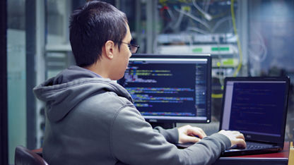 A cloud engineer sits at a desk in a server room and types on an open laptop. A desktop monitor next to the laptop displays code