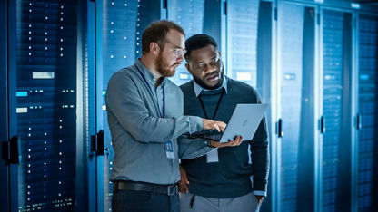 Two IT professionals stand in a server room and review information on an open laptop that one of them is holding