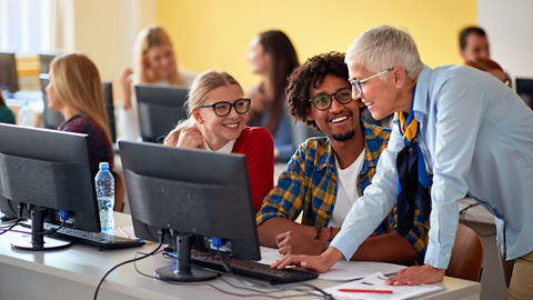 In a classroom with rows of PC workstations, a professor helps two students while looking at a monitor