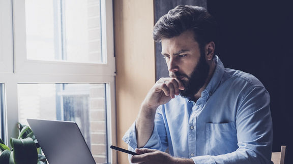 Man sitting at desk near window comparing notes in notebook on desk to content on a laptop screen
