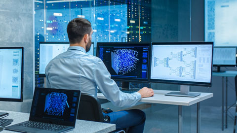 A data scientist sits at a desk in front of a glass wall in a modern data center environment reviewing data visualizations displayed across three desktop monitors. Multiple servers are visible on the other side of the glass wall