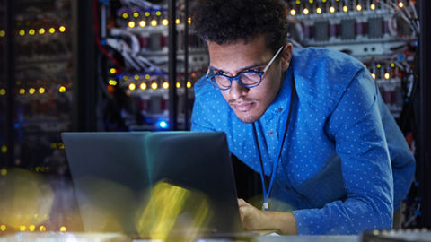 A security professional stands in a darkened server room and leans forward onto a tall surface area to work on their laptop