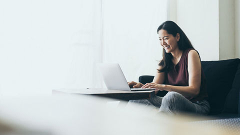 A woman sitting on a sofa smiles while typing on a laptop that rests on a coffee table