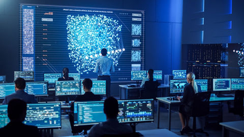 Data analysts, in a command center environment, sit at individual workstations reviewing data displayed on desktop monitors, while one person stands looking at a large data plot graphic projected on the front wall