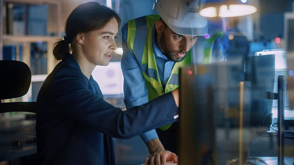 A professional woman and a man wearing a high-visibility vest look at a computer screen.