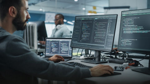 Programmer or IT professional seated in an office with two computer monitors, a laptop, and a smart phone on the desk