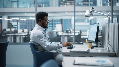 A business professional sits at a desk in a modern office space and looks at a data display on a desktop monitor while typing on a keyboard