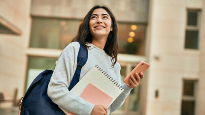 Young student smiling at something off screen, holding notebooks and a smartphone.