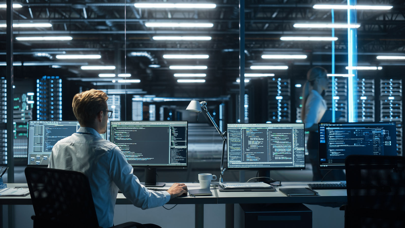 A person at a workstation surrounded by data center servers looks at code on a computer monitor