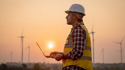 Portrait of engineer wearing yellow vest and white helmet using a computer laptop on site at wind turbines field or farm