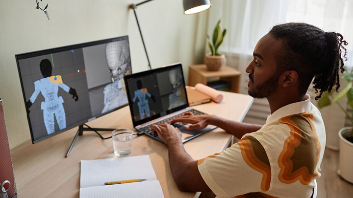 Ein Designer passt die Details der Uniform eines 3D-Charakters mit einem Laptop und einem großen Desktop-Monitor an