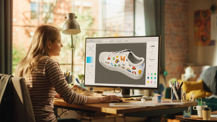 Ein Designer sitzt an einem Schreibtisch und arbeitet mit einem Desktop-Computer an dem Design eines Schuhs