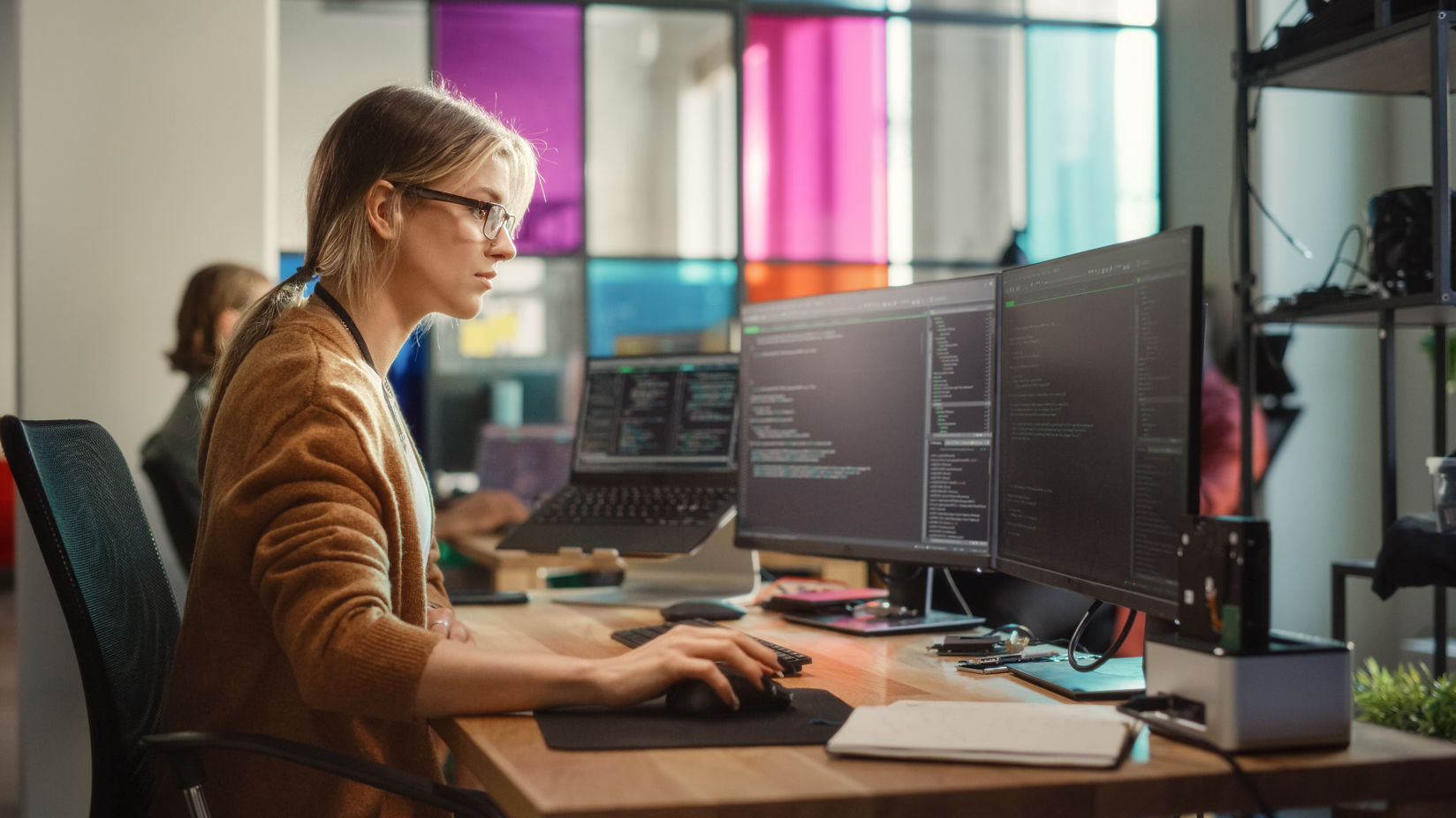 Un desarrollador sentado frente a un escritorio en un espacio de oficina moderno usando un mouse y un teclado mientras mira tres monitores en los que se observa programación.