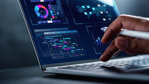 A close-up view of a person’s hand holding a pen and typing on an open laptop’s keyboard. Several multicolored graphs are shown on the screen