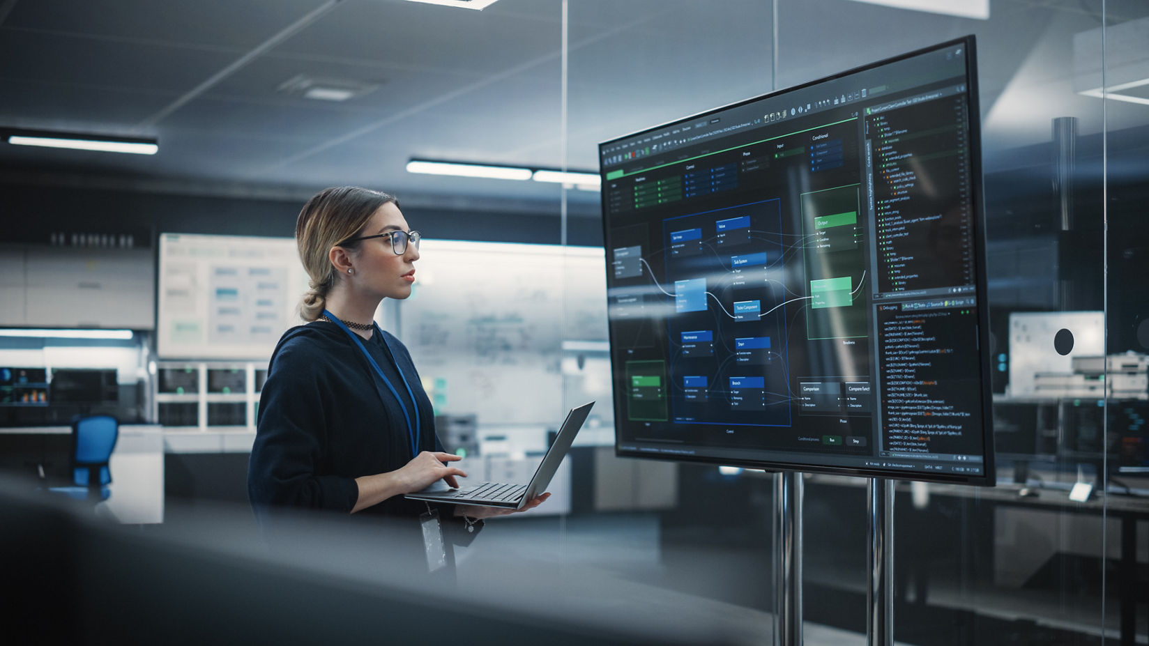 An AI professional stands in front of a wall mounted screen reviewing data displays and code in a team office environment