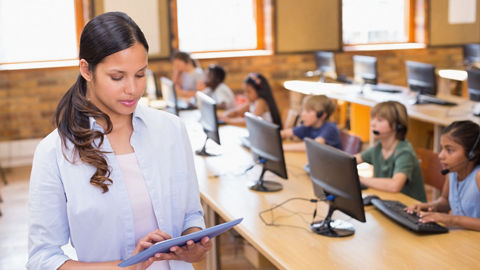 Teacher stands at front of classroom looking at a handheld tablet