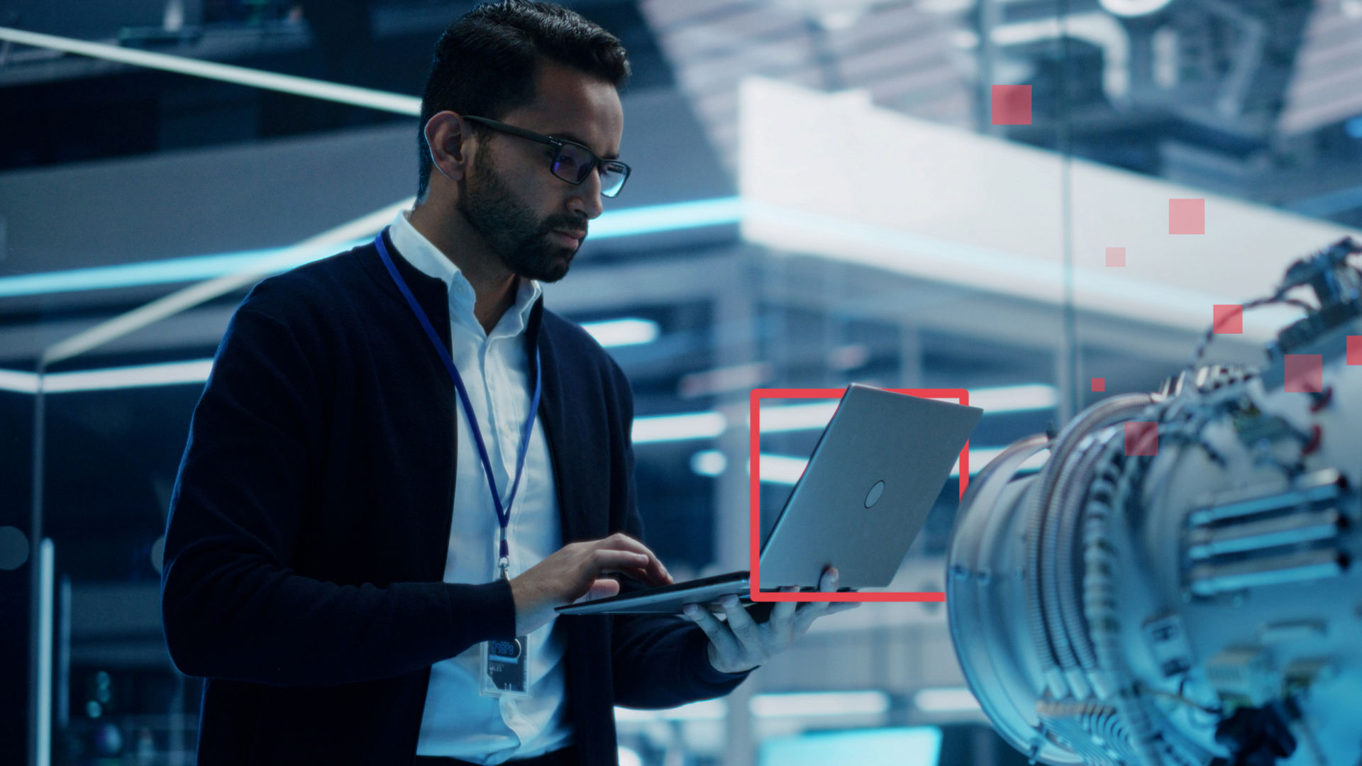 Ein Mann arbeitet in einem verglasten Büro an einem Laptop und steht in der Nähe eines großen Mikroskops; stilistische rote Quadrate heben die Technik hervor.