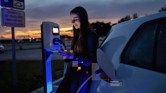 A young adult holds a cell phone while charging an electric vehicle.