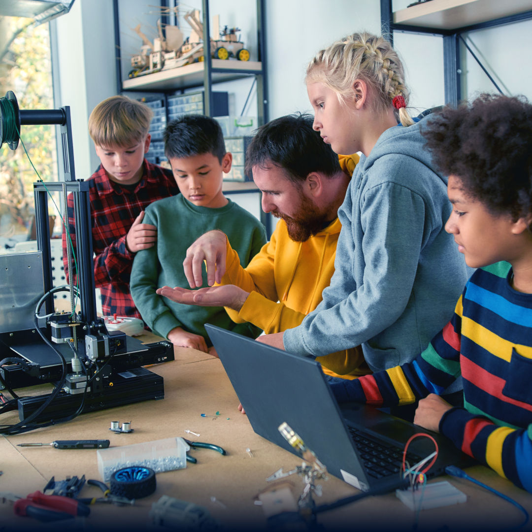 Teacher showing electronic equipment to a group of children gathered around a workbench.