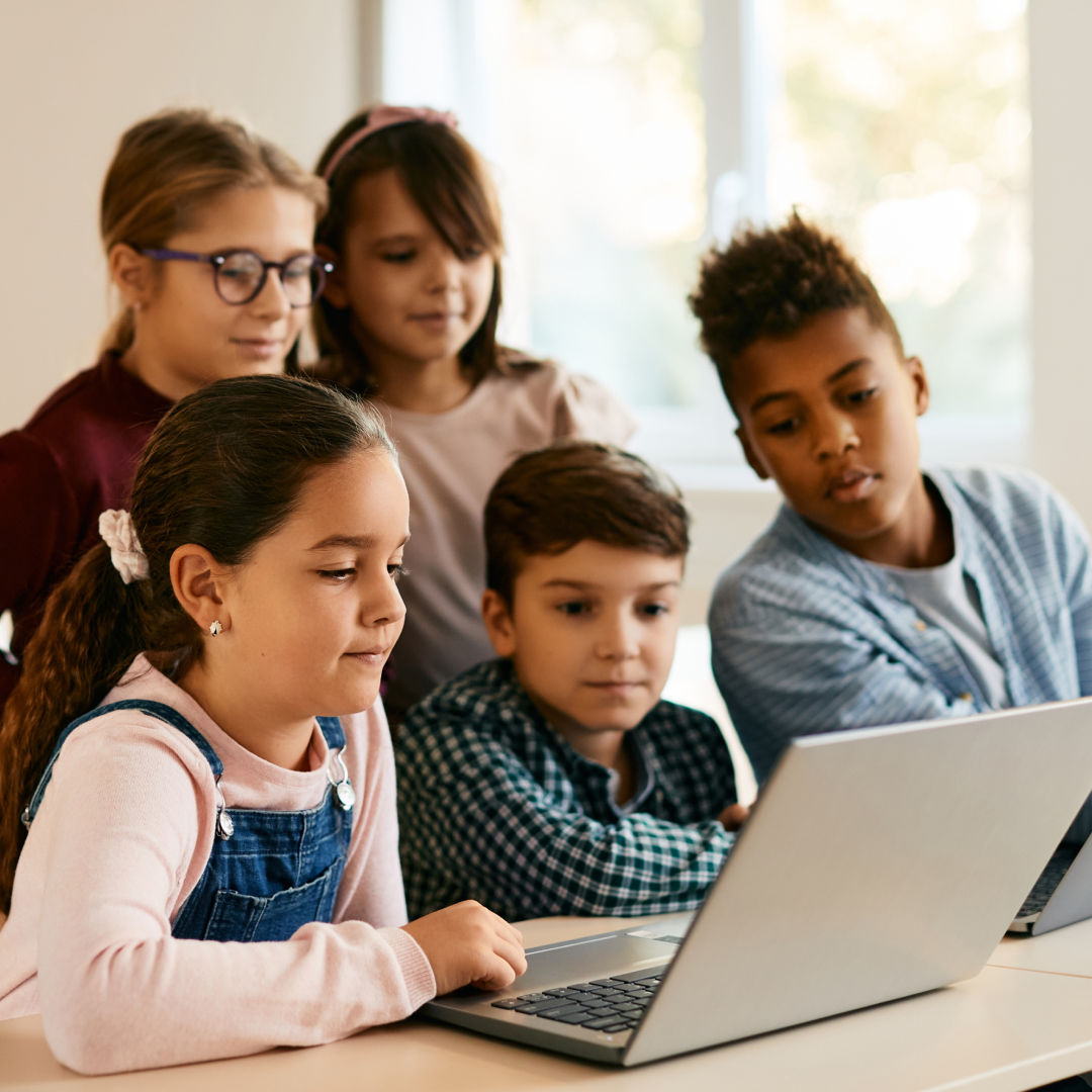 Five children gathered around a laptop at a classroom table.