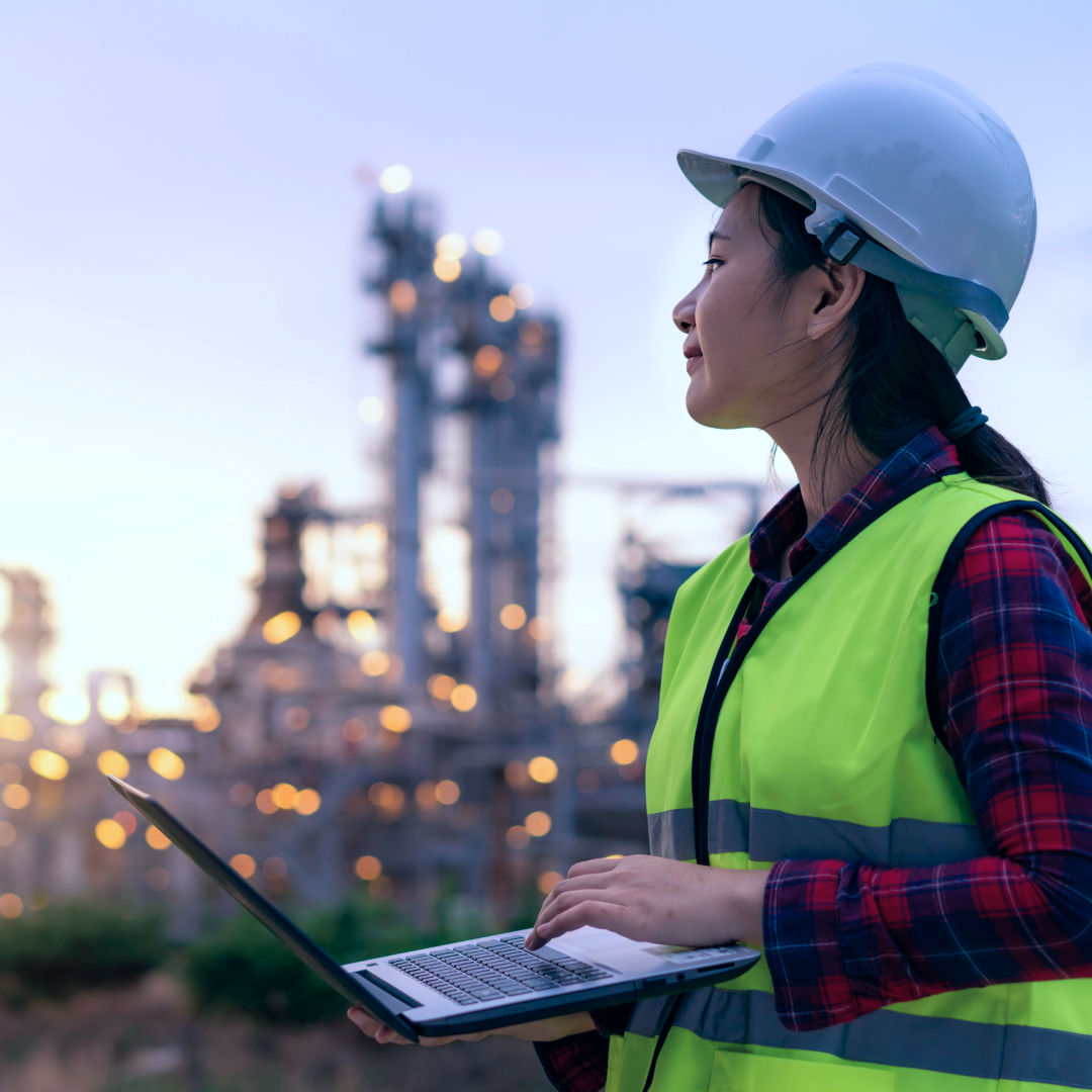 Person in a hard hat and high-visibility vest using a laptop outside an industrial facility.