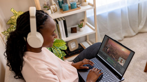 A person wearing headphones sits on a living room sofa typing on the keyboard of an open laptop resting on their legs. The frame of a movie is shown on-screen