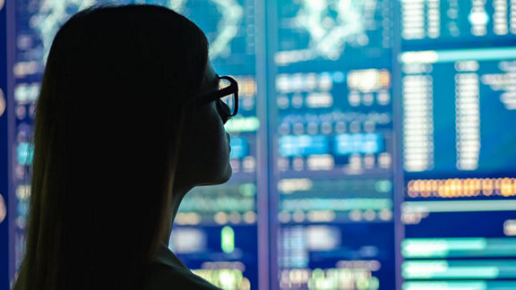 A person stands in front a wall of computer monitors that display charts and graphs