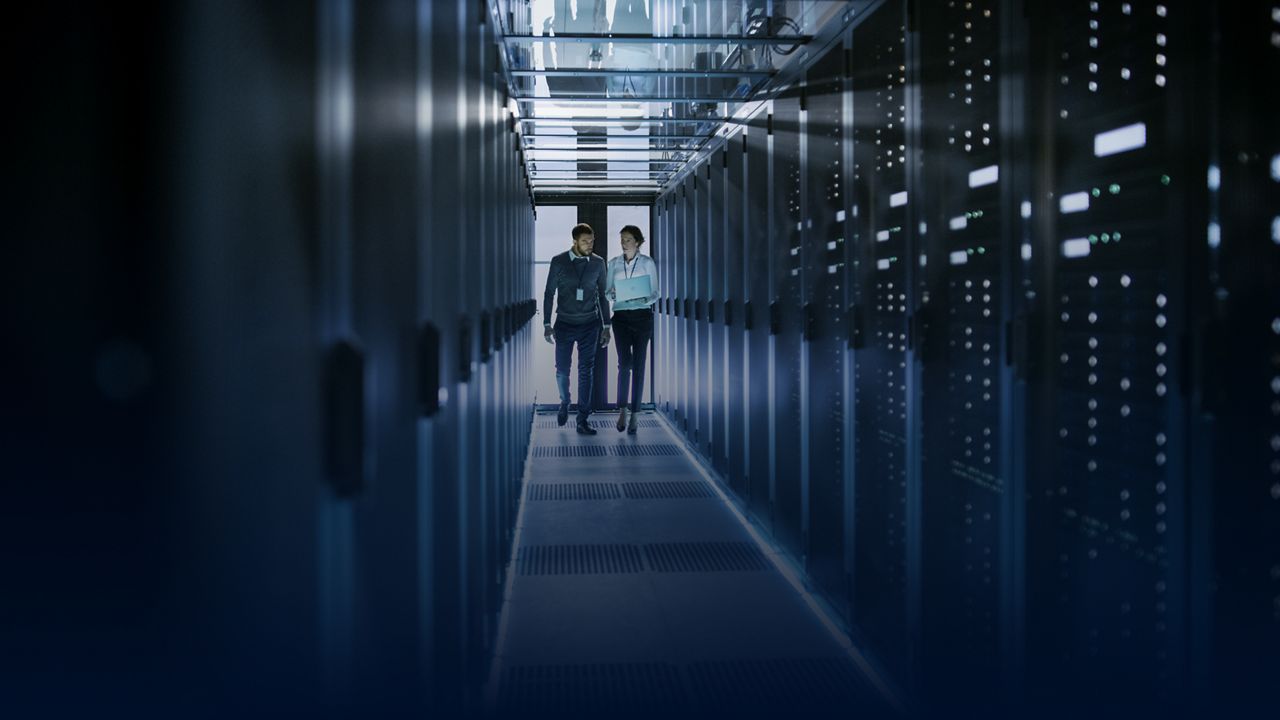 Two people walking through a data center aisle, with one holding a laptop
