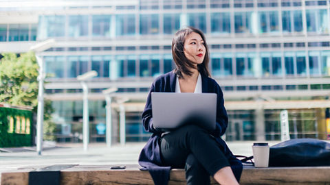 Close-up shot of a business professional sitting outside and looking off to the left in thought with an open laptop on their lap. A modern office building with many windows looms in the background