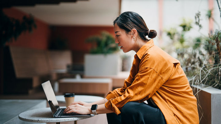 Eine Frau sitzt im Atrium eines Bürogebäudes und tippt auf einem aufgeklappten Laptop