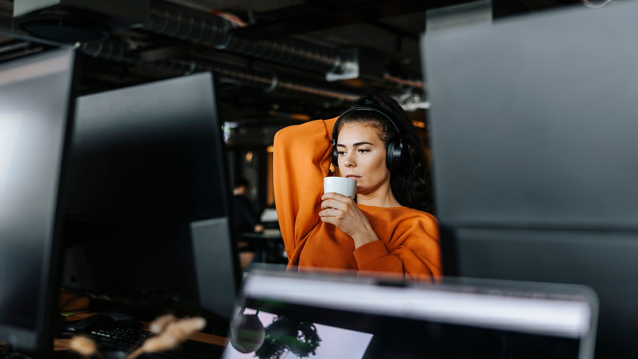 A close-up half-body view of a woman wearing headphones and an orange sweater. She is sitting in front of a desktop monitor with a cup in one hand and her other arm lifted and resting behind her head