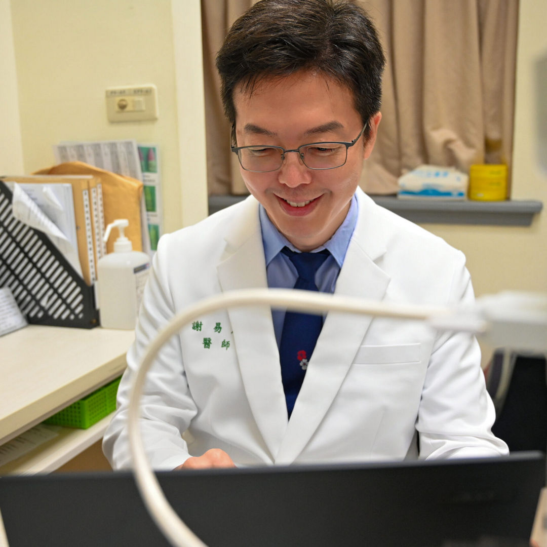 Dr. Hsieh Yi-Ting, an ophthalmologist, smiling while working on a laptop in a medical office.