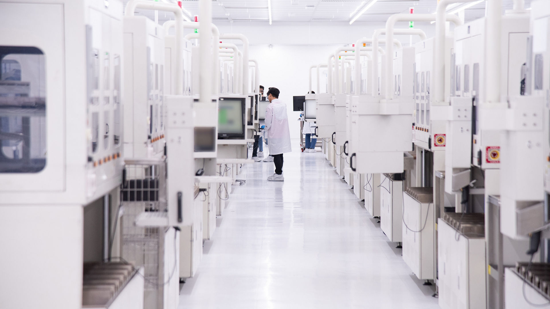A person in a white lab coat works among rows of equipment in a bright Intel assembly and packaging facility in Malaysia.