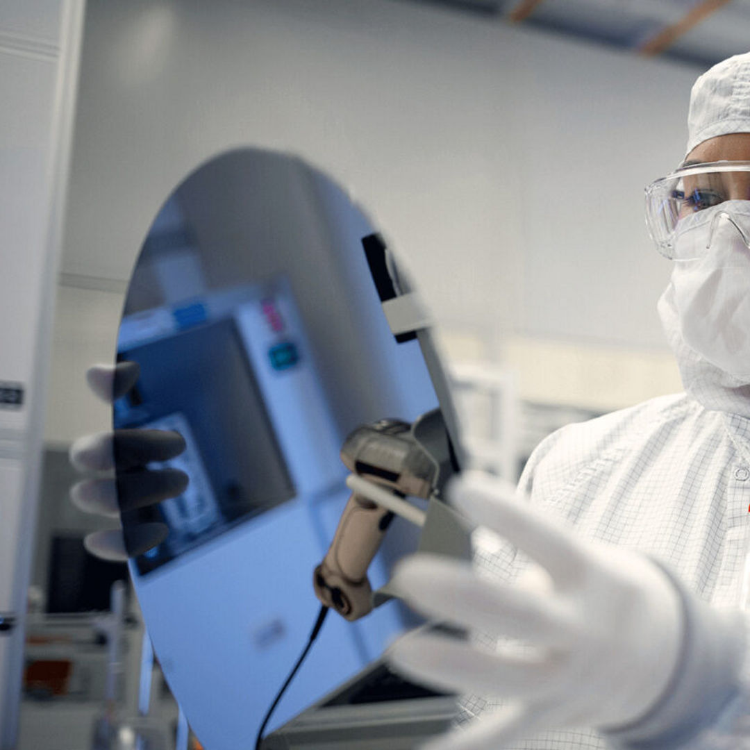 An Intel Foundry engineer in clean room gear holds a silicon waf