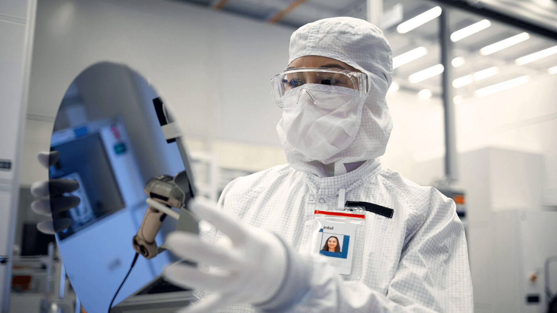 An Intel Foundry engineer in cleanroom gear holds a silicon wafer.