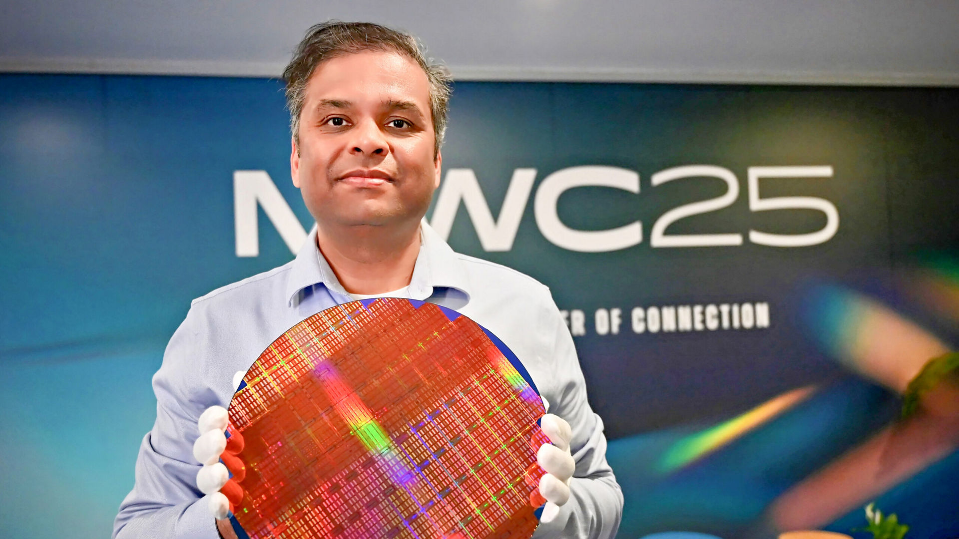 A person in a light blue shirt holds a large, circular silicon wafer with multicolored reflections. They stand in front of a blurred background with the text MWC25 The Power of Connection.