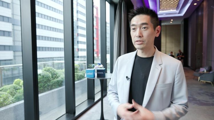 A man in a gray blazer speaks in a modern office setting.