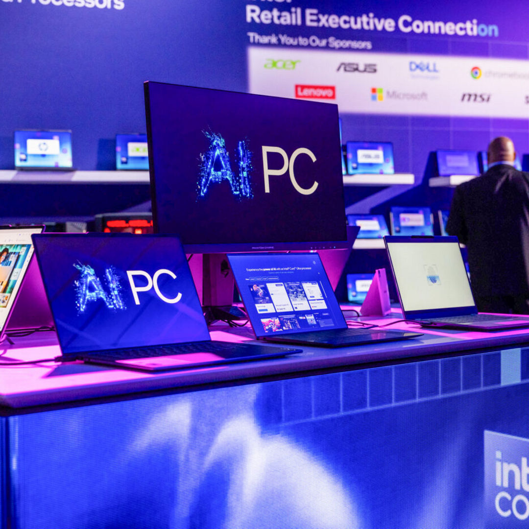 A display booth showcasing multiple laptops and monitors with AI PC graphics at a tech event. The setup is on a purple-lit table, with brand logos like Acer, ASUS, and Dell visible in the background. Attendees are in conversation nearby.