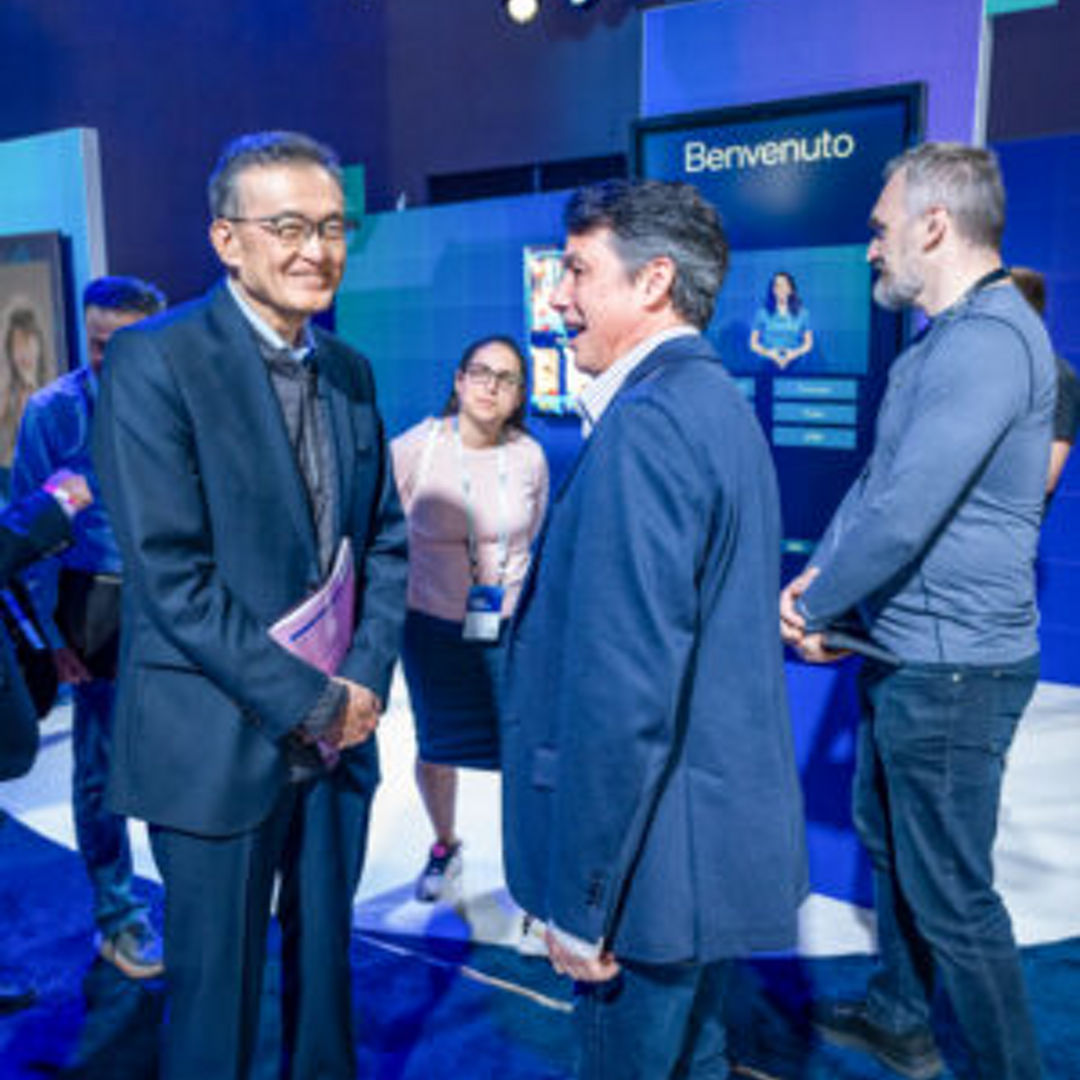 A group of people stand in a well-lit room with blue walls. Two men in suits are conversing while others watch. A screen in the background displays Benvenuto. Attendees wear name tags and appear engaged in a professional setting.