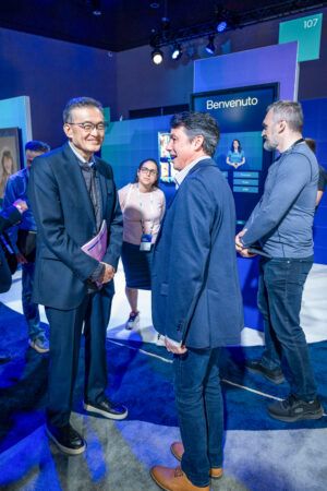 A group of people stand in a well-lit room with blue walls. Two men in suits are conversing while others watch. A screen in the background displays Benvenuto. Attendees wear name tags and appear engaged in a professional setting.