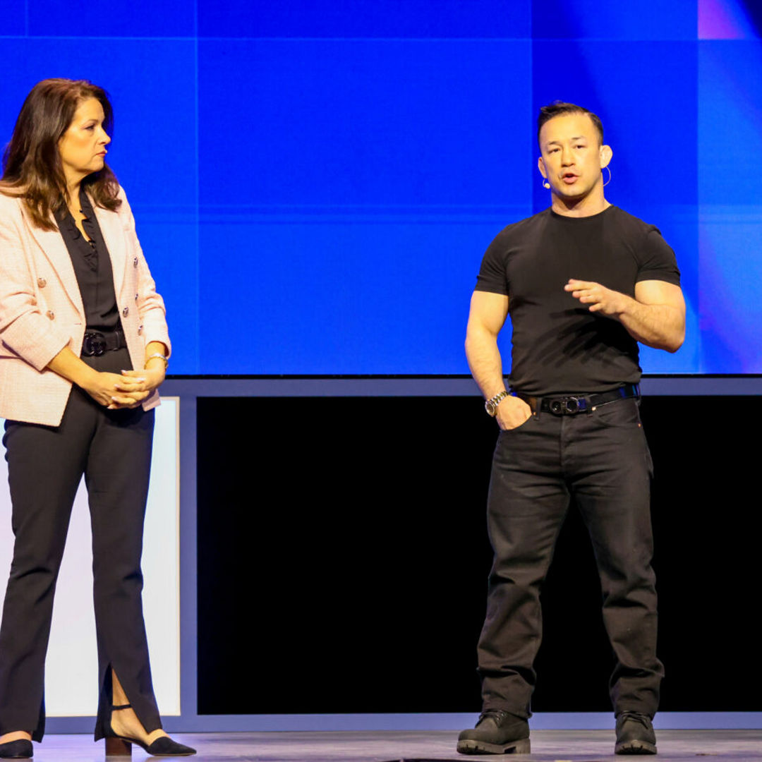 A woman in a pink blazer and dark pants stands next to a man in a black t-shirt and pants on a stage with a blue backdrop. The woman looks at him as he gestures while speaking.