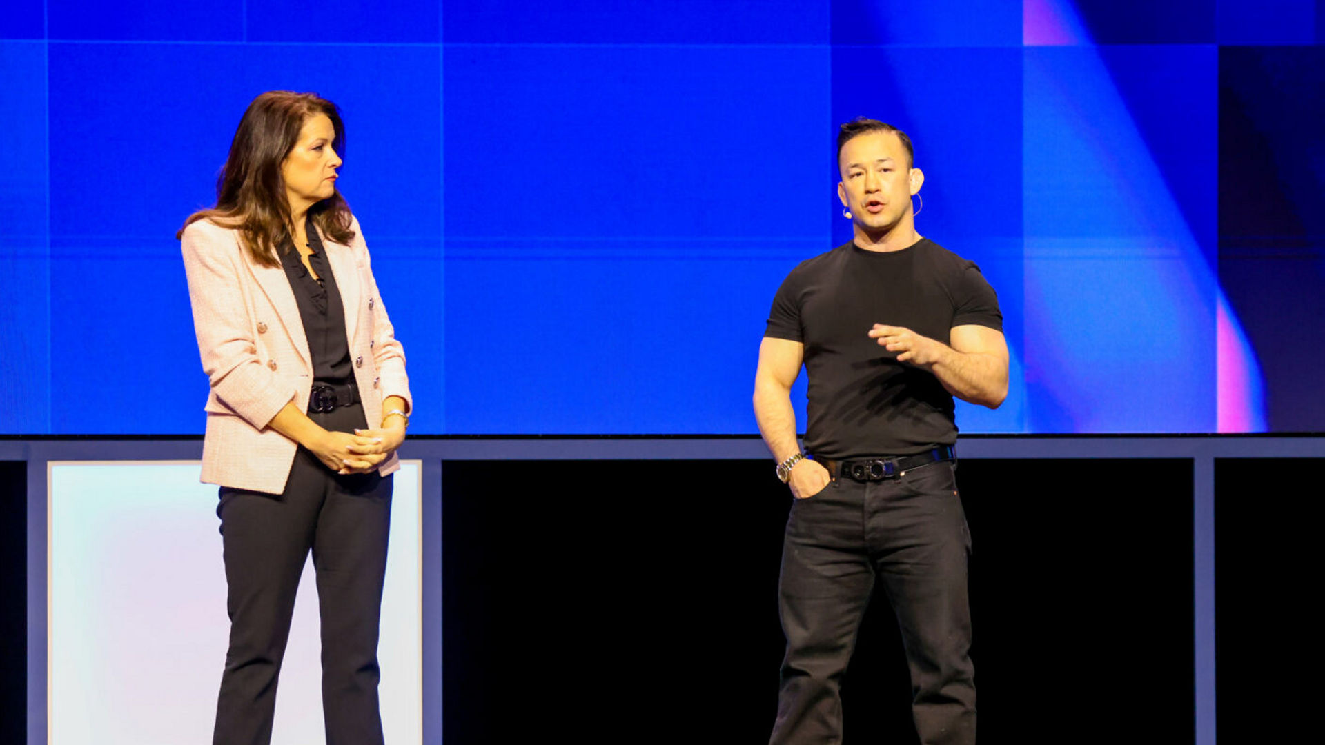 A woman in a pink blazer and dark pants stands next to a man in a black t-shirt and pants on a stage with a blue backdrop. The woman looks at him as he gestures while speaking.