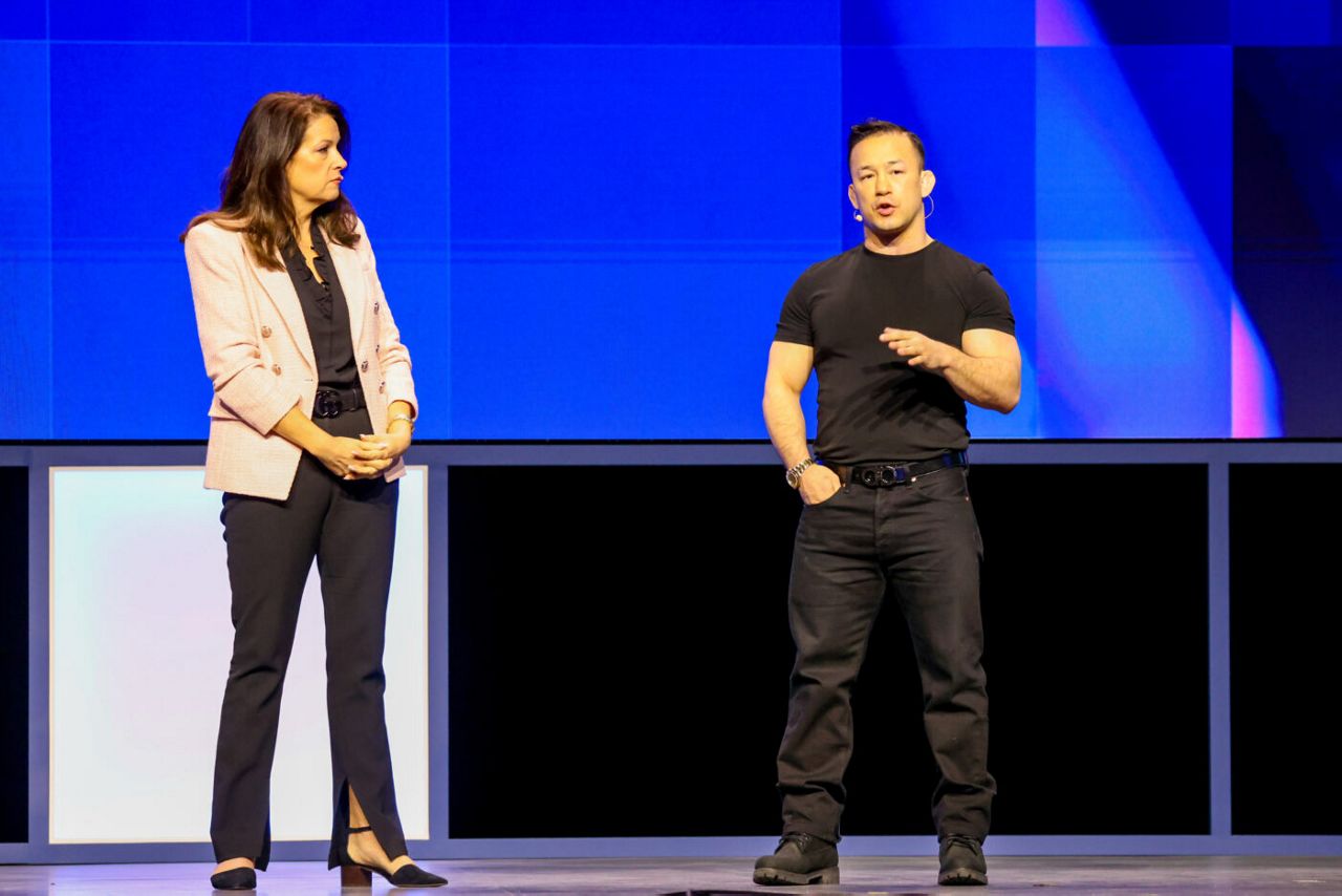 A woman in a pink blazer and dark pants stands next to a man in a black t-shirt and pants on a stage with a blue backdrop. The woman looks at him as he gestures while speaking.