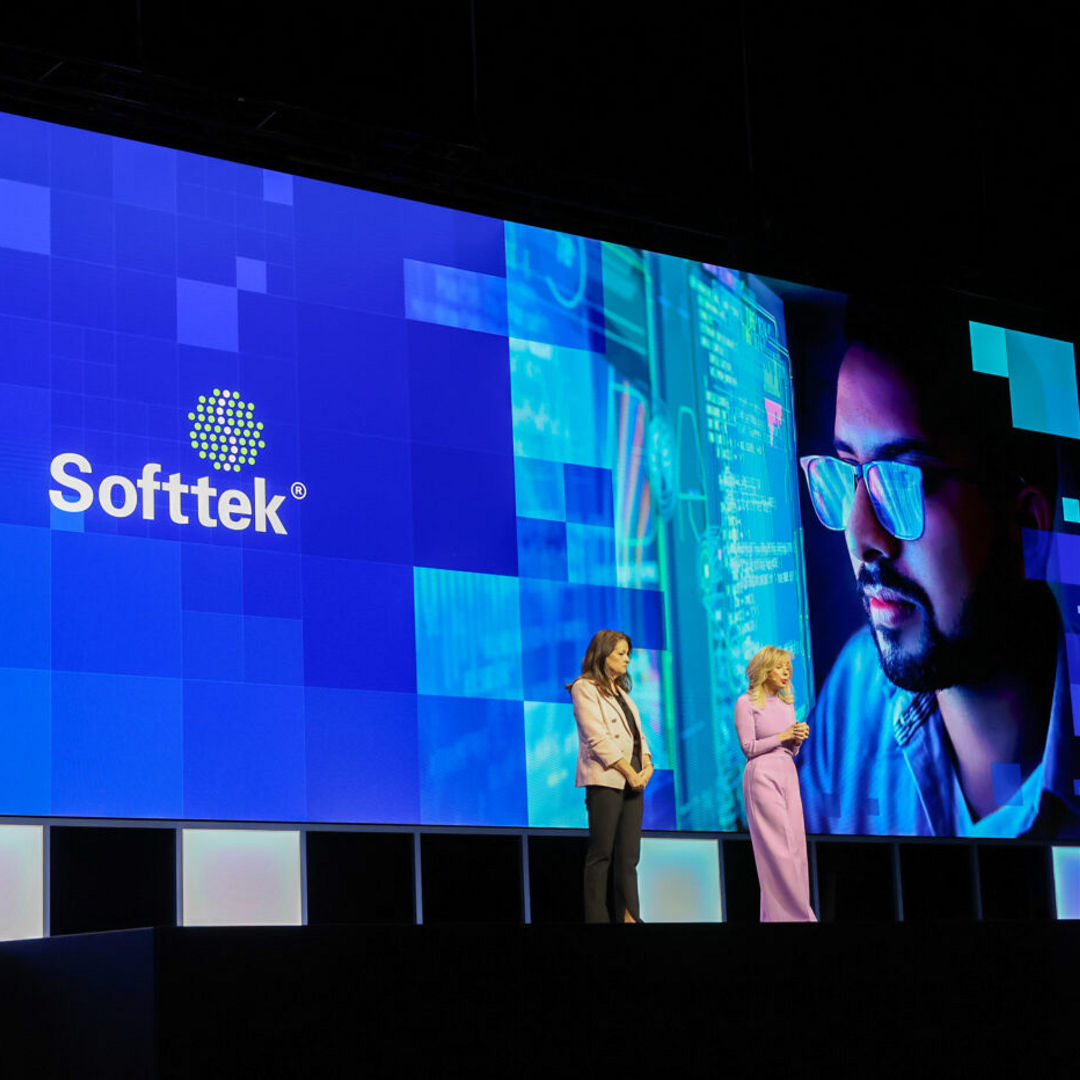 Two people stand on a stage in front of a large screen displaying the Intel and Softtek logos, along with a digitally edited image of a person wearing glasses.