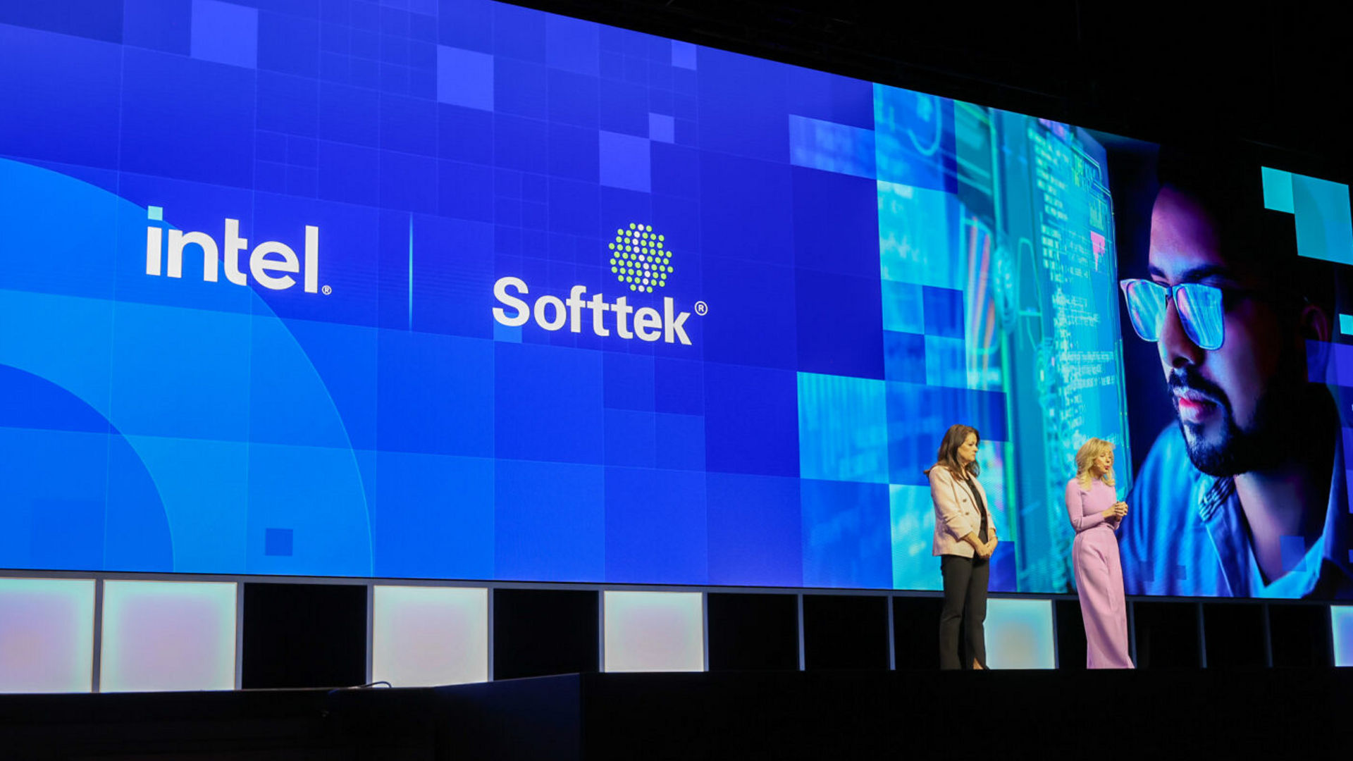 Two people stand on a stage in front of a large screen displaying the Intel and Softtek logos, along with a digitally edited image of a person wearing glasses.