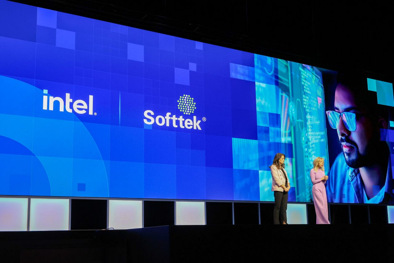 Two people stand on a stage in front of a large screen displaying the Intel and Softtek logos, along with a digitally edited image of a person wearing glasses.